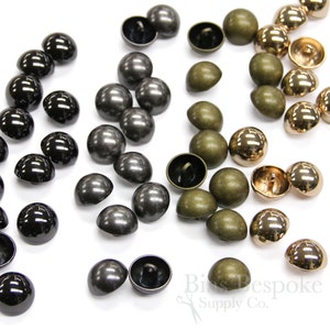 Pode incluir: Conjunto de botões em forma de cúpula em preto, prata, dourado e verde. Os botões estão dispostos em filas sobre uma superfície branca.