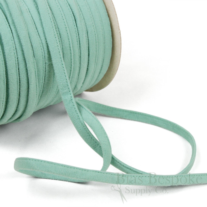 May include: A spool of light green suede cord, a type of fabric trim, is shown on a white background. The cord is rolled up on a cardboard spool. The cord is about 1/4 inch wide.