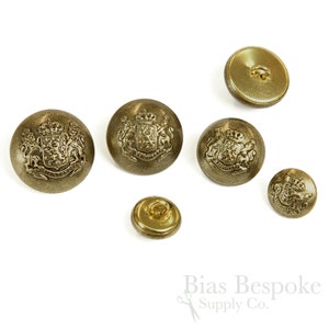 Puede incluir: Seis botones de color dorado con un diseño en relieve de un escudo de armas con una corona y dos leones. Los botones están dispuestos en semicírculo sobre un fondo blanco.