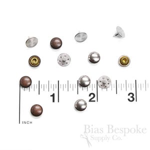 May include: Assortment of 10 metal rivets in silver and brown colors. The rivets are 1/2 inch in diameter and are shown on a ruler for size reference. Bias Bespoke Supply Co. is printed on the bottom of the image.