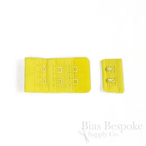 May include: A bright yellow bra extender featuring three columns of metal hooks and eyes on the larger piece, and a matching smaller piece with two rows of metal loops. The fabric is a textured, woven material with a consistent vibrant yellow hue.