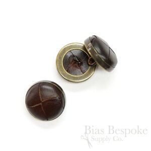 May include: Three brown leather buttons with a brass rim. The buttons have a cross-stitch pattern on the leather.