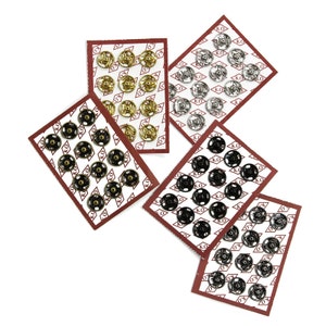 May include: Four cards of metal snap fasteners, each card has a different color of fasteners. The colors are gold, silver, black, and nickel. The cards are all on a white background.