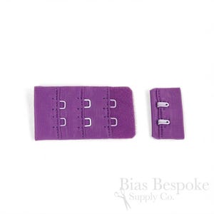 May include: A purple bra extender featuring three columns of hook-and-eye closures on the left piece and two rows of eyes on the right piece. The fabric is a solid purple textile with matching stitching and silver-toned metal hardware components.