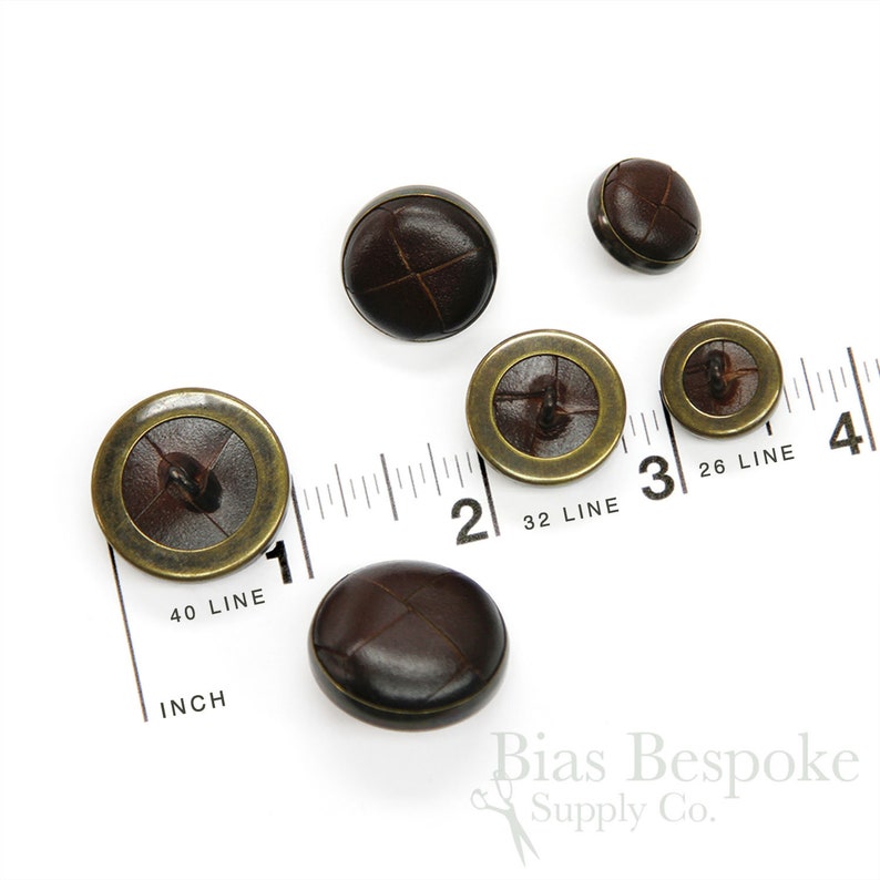 May include: Five brown leather buttons with a brass rim. The buttons are arranged on a white surface with a ruler in the background. The ruler is marked in inches and lines. The text on the ruler reads "40 LINE", "INCH", "32 LINE", "26 LINE".