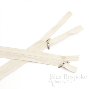 Può includere: Due nastri di cerniera beige con tiretti in metallo. I nastri di cerniera sono disposti a croce. Bias Bespoke Supply Co. è stampato in nero su sfondo bianco nella parte inferiore dell'immagine.