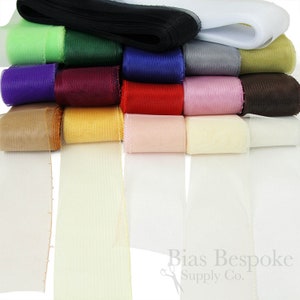 May include: A collection of 15 rolls of tulle ribbon in various colors, including white, black, green, red, brown, purple, and yellow. The ribbon is arranged in a stack, with the rolls of ribbon unfurling at the bottom of the image.