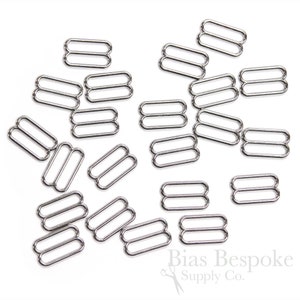 May include: A collection of silver-colored metal bra strap sliders, each with a double-loop design. The hardware is scattered across a white surface, suitable for lingerie and sewing projects.