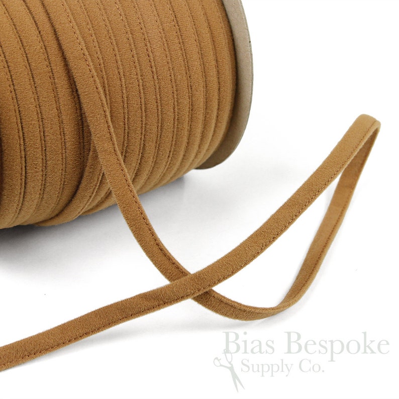 May include: A spool of brown cord or ribbon with a soft, suede-like texture. The cord is approximately 1/4 inch wide and is wound around a cardboard spool.