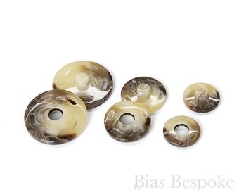 以下が含まれることがあります： 茶色とクリーム色のプラスチック製のホックが5個セットになっています。ボタンは丸型で、中央が盛り上がっています。白い背景に半円形に配置されています。
