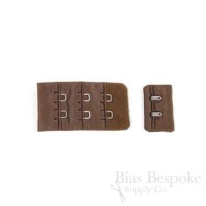 May include: A brown bra extender featuring three columns of hook-and-eye closures on a fabric panel, accompanied by a matching smaller fabric piece with two eye loops. The components are made of a textured brown material with metal hardware.