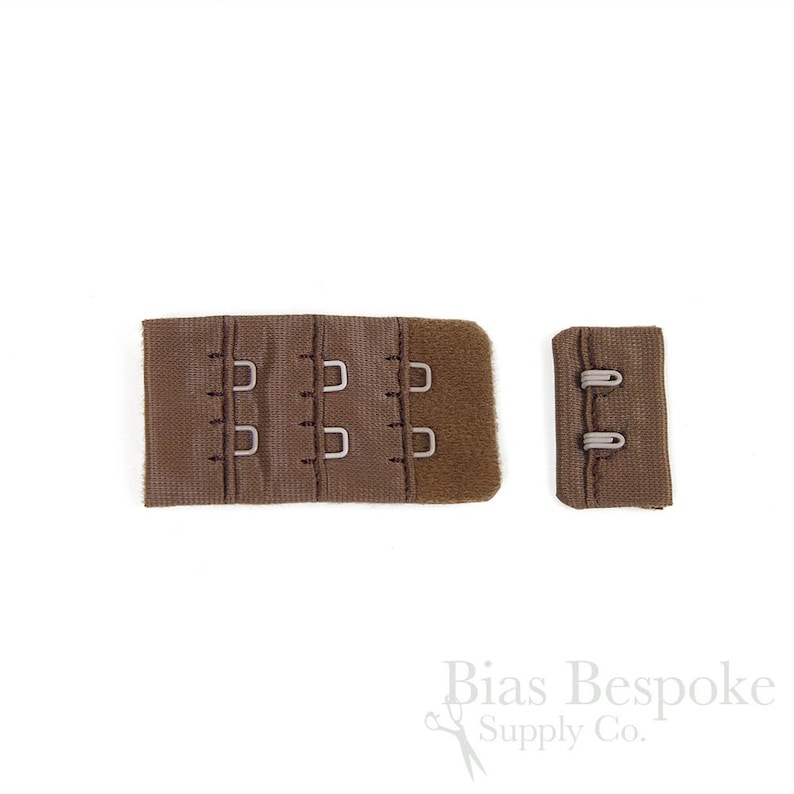 May include: A brown bra extender featuring three columns of hook-and-eye closures on a fabric panel, accompanied by a matching smaller fabric piece with two eye loops. The components are made of a textured brown material with metal hardware.