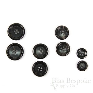 Puede incluir: Conjunto de ocho botones de cuerno negro con cuatro agujeros cada uno. Los botones están dispuestos en círculo sobre un fondo blanco.