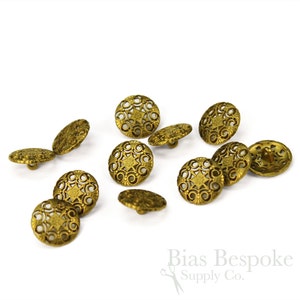 Può includere: Dieci bottoni in metallo color oro con un design decorativo e intricato. I bottoni hanno una forma circolare con un centro a forma di stella rialzato.