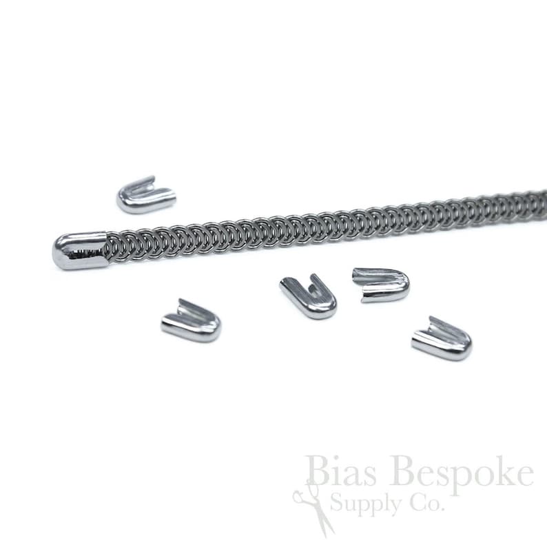 May include: Close-up of a silver-colored chain with a rounded end and several U-shaped metal clasps scattered around it. The chain is made of small, interconnected rings. The clasps are shiny and reflect the light. The background is white.