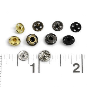 以下が含まれることがあります： 金、銀、黒、ブロンズなど、さまざまな仕上げの金属製スナップボタン10個の詰め合わせ。ボタンの直径は約2.5cmで、中央が盛り上がっています。