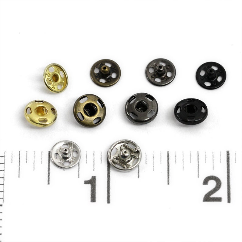 以下が含まれることがあります： 金、銀、黒、ブロンズなど、さまざまな仕上げの金属製スナップボタン10個の詰め合わせ。ボタンの直径は約2.5cmで、中央が盛り上がっています。