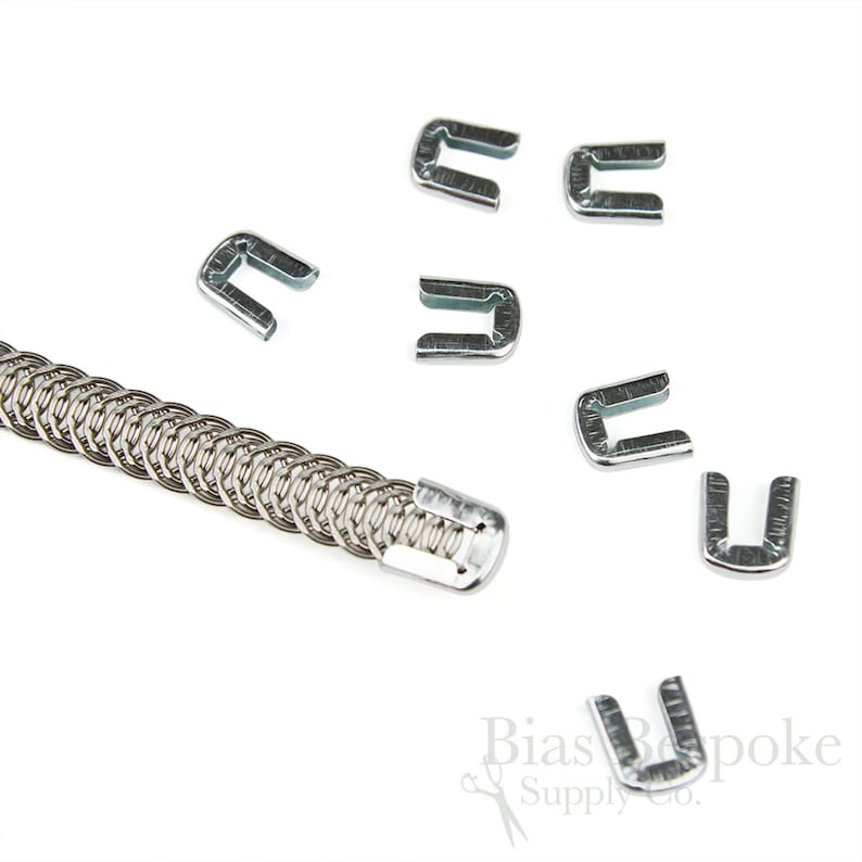May include: A collection of silver-colored metal U-shaped clasps and a chainmail-style metal tube. The clasps are scattered around the tube, all on a white background. The image is well-lit, highlighting the metallic sheen of the objects.