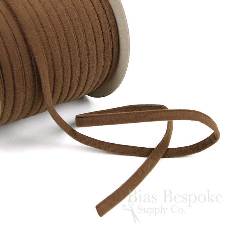 May include: A spool of brown bias tape, a type of fabric trim, is shown on a white background. The tape is rolled up on a cardboard spool and a length of the tape is extended out to the right. The tape is a soft, brown color and has a slightly textured surface.