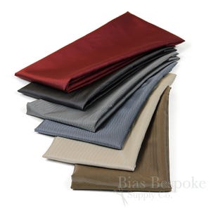 May include: A stack of six fabric samples in various colors and herringbone patterns. The colors are burgundy, black, gray, blue, beige, and brown. The fabric is smooth and has a subtle sheen. Bias Bespoke Supply Co. is printed on the bottom fabric sample.