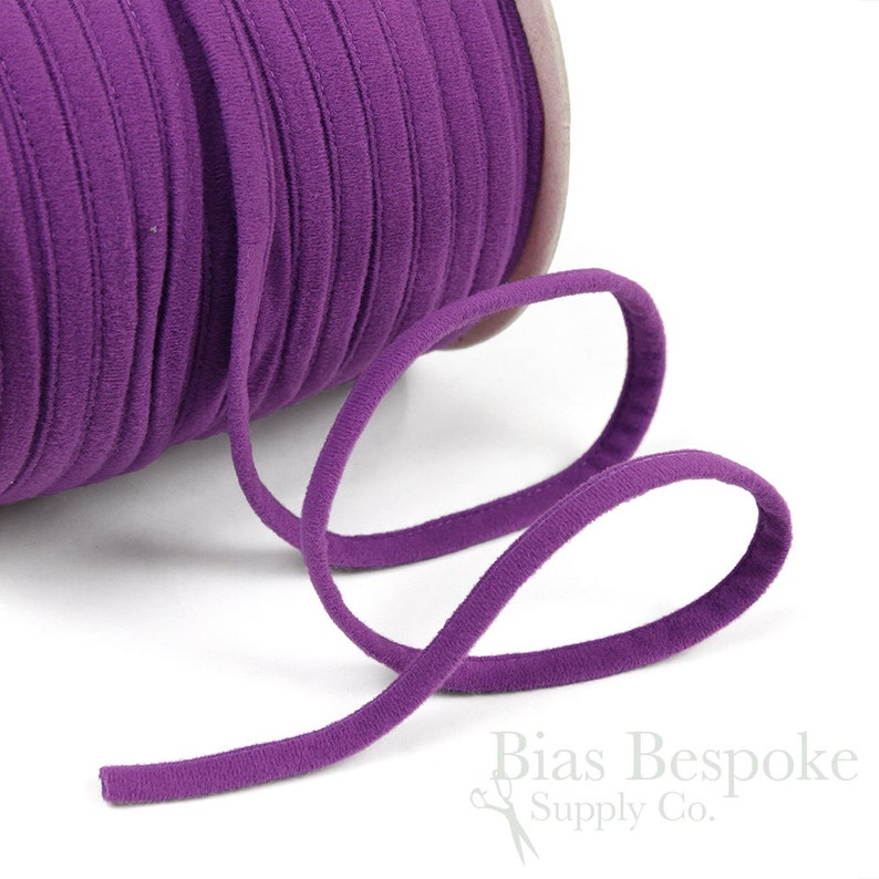 May include: A spool of purple elastic cord, also known as bias tape, is shown on a white background. The cord is soft and flexible, and it is perfect for sewing projects.