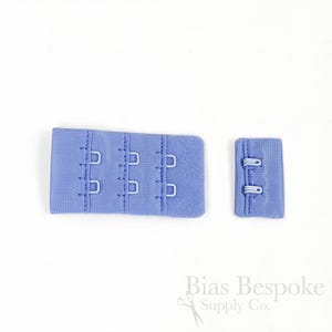 May include: A light blue bra extender featuring three rows of metal hooks and eyes on a fabric panel, accompanied by a matching smaller fabric panel with two metal loops. The text Bias Bespoke Supply Co. is visible in the bottom right corner.