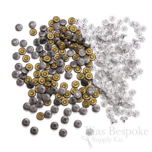 Op de afbeelding: Een grote verzameling metalen drukknopen in zilver en antiek messing. De drukknopen zijn verspreid over een witte achtergrond.