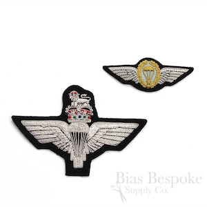 May include: Two embroidered patches, one silver and one gold, with a parachute design. The silver patch features a crown and wings, while the gold patch has a wreath around the parachute. Both patches are on a black background.