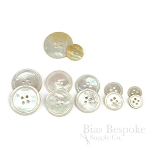 May include: A collection of iridescent, pearlescent buttons in various sizes. The buttons have four holes and a subtle, shimmering quality. The buttons are arranged on a white background, showcasing their different sizes and shapes.
