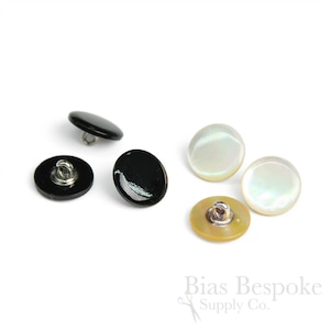Puede incluir: Una colección de botones redondos en negro y blanco iridiscente. Los botones negros tienen un acabado brillante, mientras que los botones blancos tienen un brillo nacarado. Cada botón tiene un vástago de metal para coser.