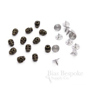 Könnte beinhalten: Ein Set mit 14 antiken goldenen Totenkopf-Knöpfen und 8 silbernen Nieten-Sets. Die Totenkopf-Knöpfe haben einen Durchmesser von etwa 1 cm. Die Nieten-Sets haben einen Durchmesser von etwa 1,5 cm. Die Nieten-Sets haben eine silberne Oberfläche und eine spitze Spitze. Die Totenkopf-Knöpfe sind perfekt, um Ihren Kleidungsstücken oder Accessoires einen Hauch von Gothic-Stil zu verleihen.