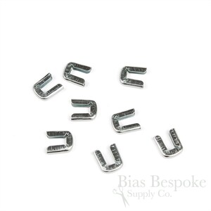 May include: A collection of silver-colored metal U-shaped clasps, scattered on a white background. The clasps are small, with a rectangular shape and a curved top. The image is well-lit, highlighting the metallic sheen of the clasps.