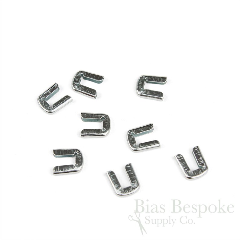 May include: A collection of silver-colored metal U-shaped clasps, scattered on a white background. The clasps are small, with a rectangular shape and a curved top. The image is well-lit, highlighting the metallic sheen of the clasps.