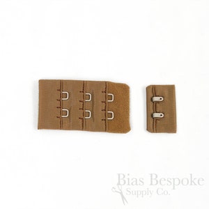 May include: A tan bra extender featuring three rows of metal hooks and eyes on a fabric base, accompanied by a separate matching two-hook closure piece. The text Bias Bespoke Supply Co. is printed in the bottom right corner of the image.