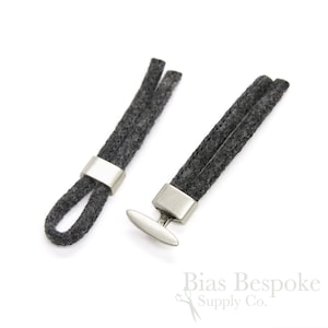 Puede incluir: Dos extremos de cordón de fieltro gris con botones de metal plateado. Un botón es un simple bucle, el otro es una barra en forma de T.