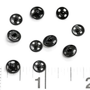 以下が含まれることがあります： 白い表面に、ポッパーとも呼ばれる黒い金属製のスナップファスナーが10個散らばっています。画像の下部には定規が見えます。