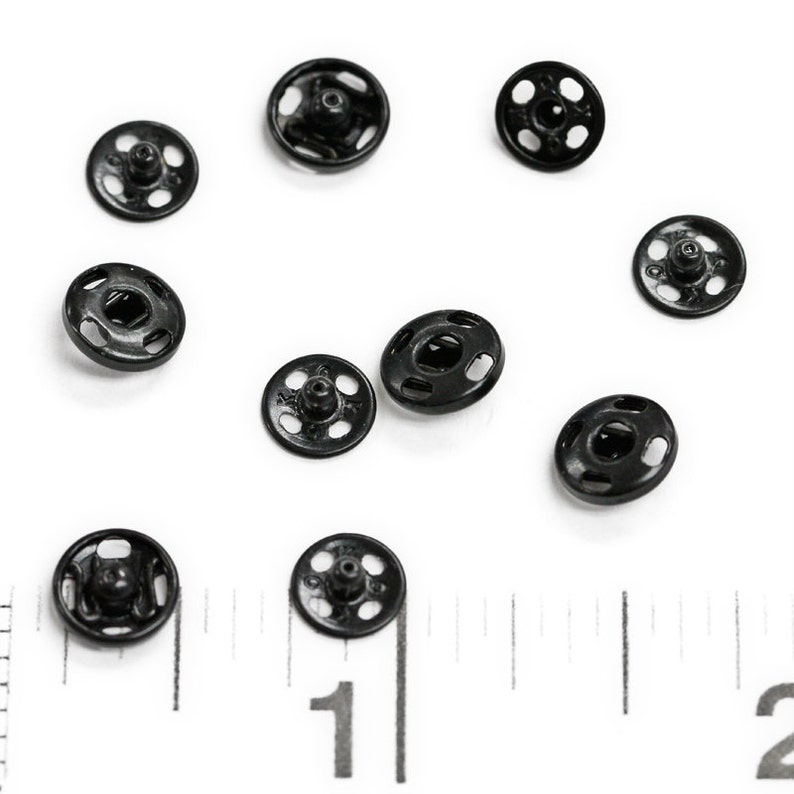 以下が含まれることがあります： 白い表面に、ポッパーとも呼ばれる黒い金属製のスナップファスナーが10個散らばっています。画像の下部には定規が見えます。