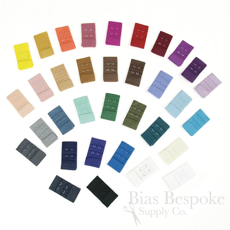 May include: A collection of rectangular bra hook and eye closures in a wide variety of colors, including yellow, orange, pink, red, brown, green, blue, purple, white, and black. Each piece features three rows of metal hooks and eyes.