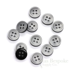 May include: A collection of 11 gray plastic buttons with four holes each, and one black plastic button with four holes. The buttons are arranged in a circle on a white background.