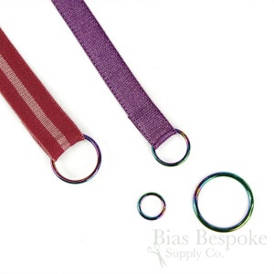 Könnte beinhalten: Zwei Stoffbänder mit unterschiedlichen Mustern, eines rot mit weißen Streifen und eines lila, jeweils an einem silberfarbenen Metallring befestigt. Drei weitere silberfarbene Metallringe in verschiedenen Größen sind ebenfalls zu sehen. Der Text "Bias Bespoke Supply Co." ist in der rechten unteren Ecke des Bildes sichtbar.