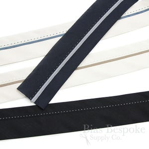 May include: Close-up of several bias tape strips in various colors. The strips include navy blue, black, and off-white, each with a contrasting stripe and decorative stitching. The Bias Bespoke Supply Co. logo is visible in the bottom right corner.