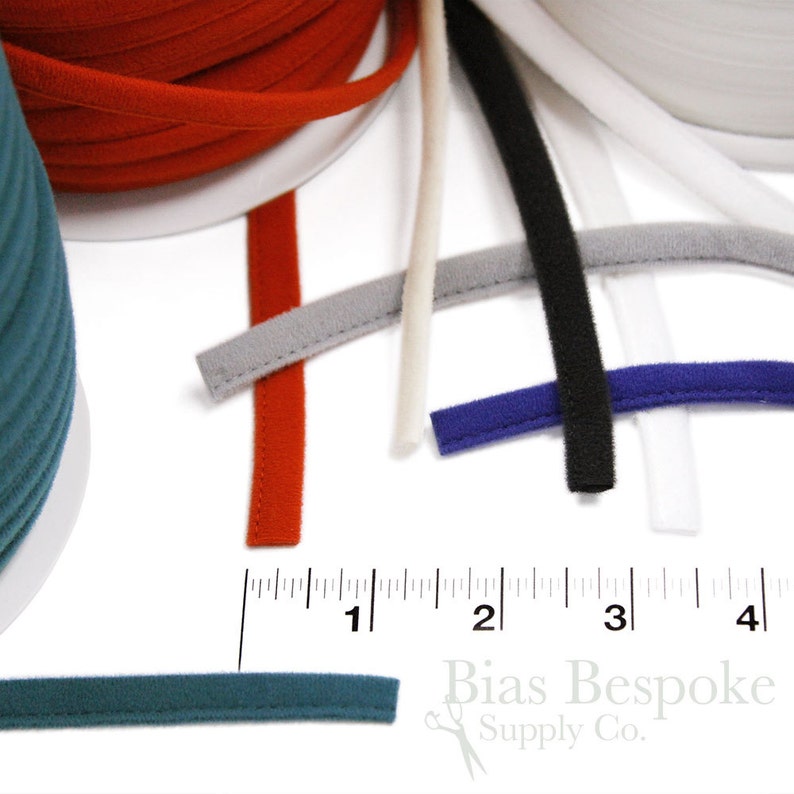 May include: A close-up of various colors of 1/4 inch wide bias tape, including red, white, black, gray, blue, and teal. The tape is laid out on a white surface with a ruler showing the measurement of 1 inch.