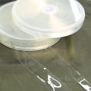May include: Two rolls of clear, transparent bias tape are stacked on top of each other. The tape is unwound from the rolls, with two strips extending towards the bottom of the image. The rolls are in a clear plastic container.