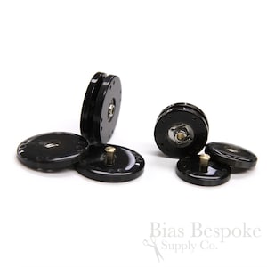May include: A collection of black magnetic snaps with a shiny finish. Each snap has a circular design with small holes around the edge and a central metal component. The snaps are designed for use in sewing and crafting projects.