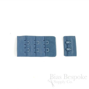 May include: A blue bra hook and eye closure set consisting of a wide fabric strip with three columns of metal loops and a narrower fabric strip featuring two metal hooks. The components are made of a textured blue material with matching metal hardware.