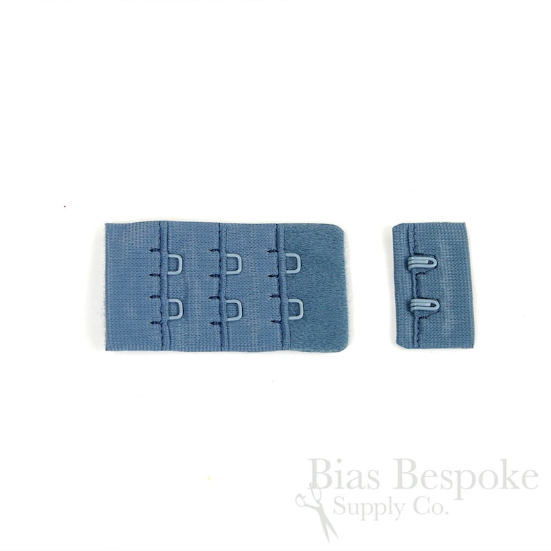 May include: A blue bra hook and eye closure set consisting of a wide fabric strip with three columns of metal loops and a narrower fabric strip featuring two metal hooks. The components are made of a textured blue material with matching metal hardware.