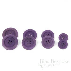 Puede incluir: Conjunto de seis botones de madera de color morado, cada uno con cuatro agujeros. Los botones están dispuestos en dos filas de tres, con los botones más grandes en la parte posterior y los botones más pequeños en la parte delantera.