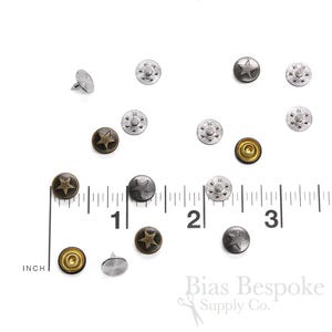 Peut inclure: Assortiment de boutons-pression en métal en forme d'étoile, en argent, or et noir. Les boutons-pression sont dispersés sur une surface blanche avec une règle en arrière-plan. La règle est graduée en pouces.