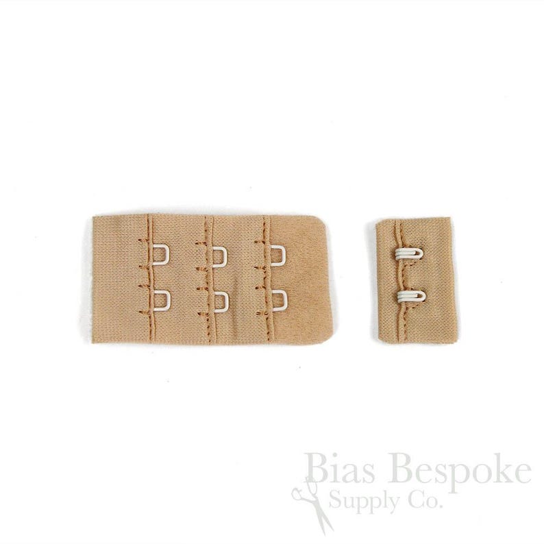 May include: A beige bra extender set featuring a wide panel with three columns of metal hooks and a corresponding narrow panel with two rows of metal eyes. The fabric is a soft, neutral tan color with visible stitching along the edges and fasteners.