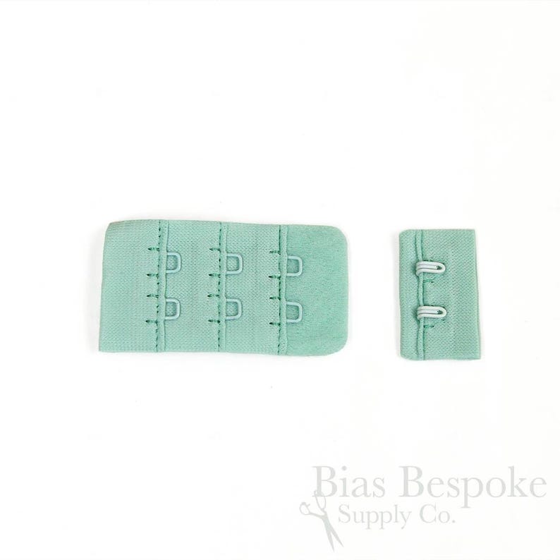 May include: A mint green bra extender featuring three columns of metal hooks and eyes on a fabric base, accompanied by a smaller matching fabric piece with two metal loops. The text Bias Bespoke Supply Co. is printed in the bottom right corner.